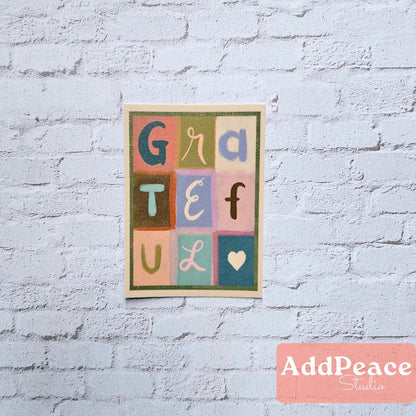 Colorful sticker with the word 'GRATEFUL' on a light blue brick wall, featuring the brand 'AddPeace Studios'.