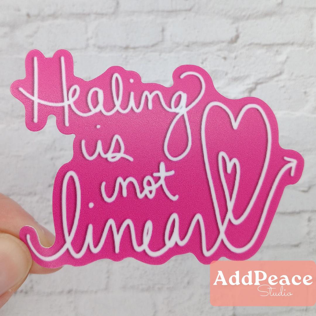 Pink sticker with 'Healing is not linear' text held by a hand on a textured white background, with 'AddPeace Studio' branding.