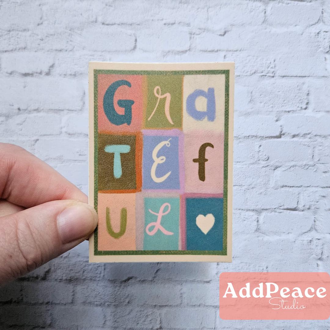 Hand holding a sticker with colorful letters spelling 'GRATEFUL' against a white brick wall.
