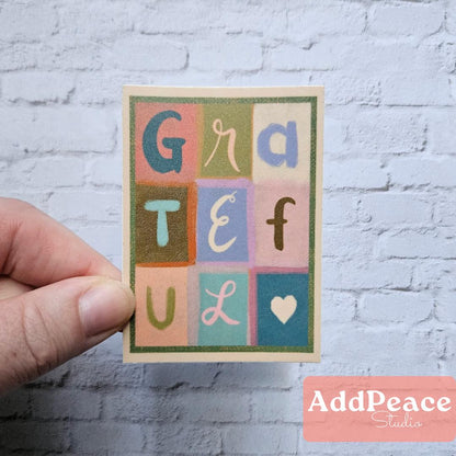 Hand holding a sticker with colorful letters spelling 'GRATEFUL' against a white brick wall.